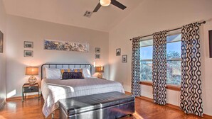 A bedroom with a bed, bedside tables, a ceiling fan, a window with patterned curtains, and a view of the outdoors.