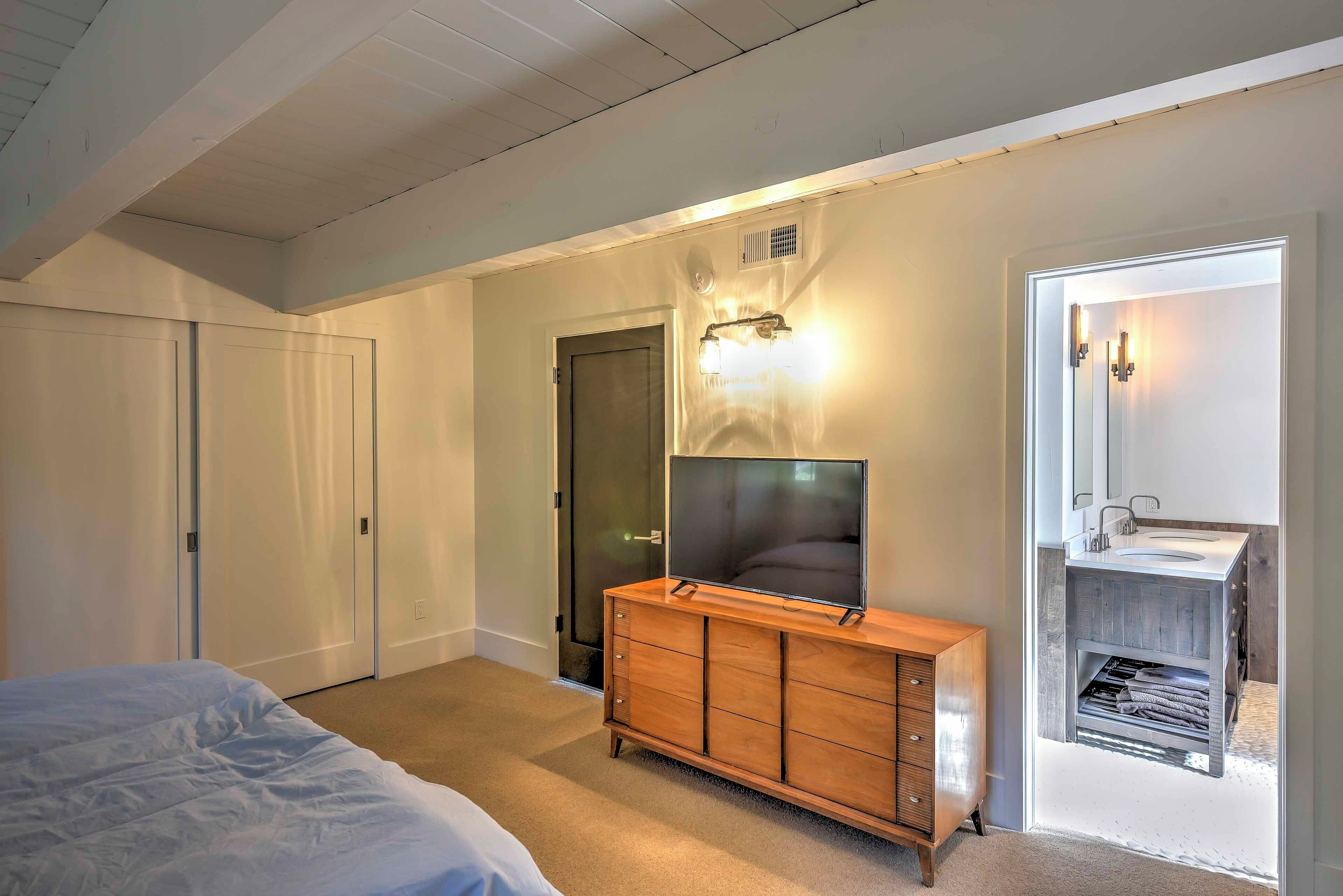 Une chambre à coucher comprenant un lit, une commode en bois, un téléviseur, une salle de bain avec un lavabo et un porte-serviettes, et une porte donnant sur une autre pièce.