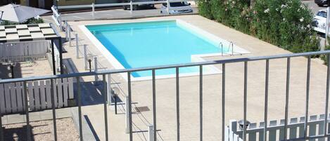 Une piscine entourée d’une clôture blanche, un parasol à carreaux et un bâtiment visible au loin.