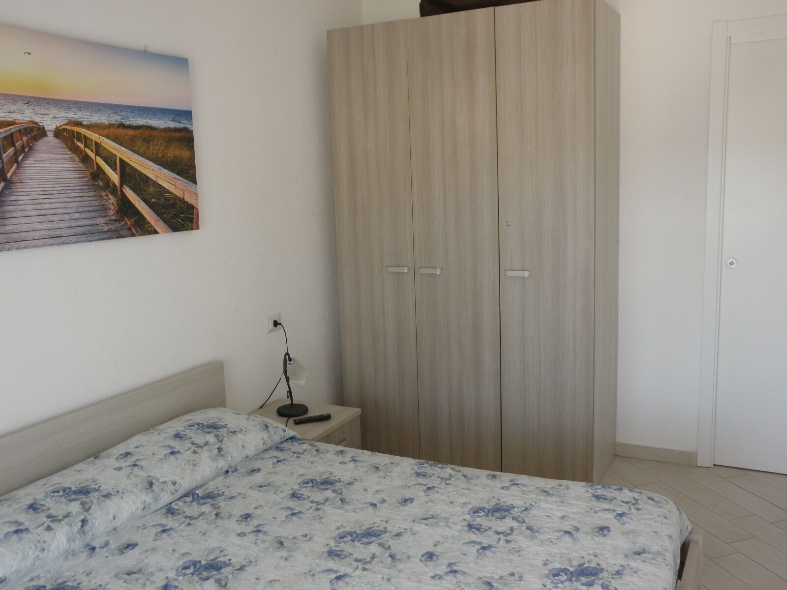 Une chambre à coucher comprenant un lit, une armoire, une table de chevet avec une lampe et un tableau représentant une scène de plage.