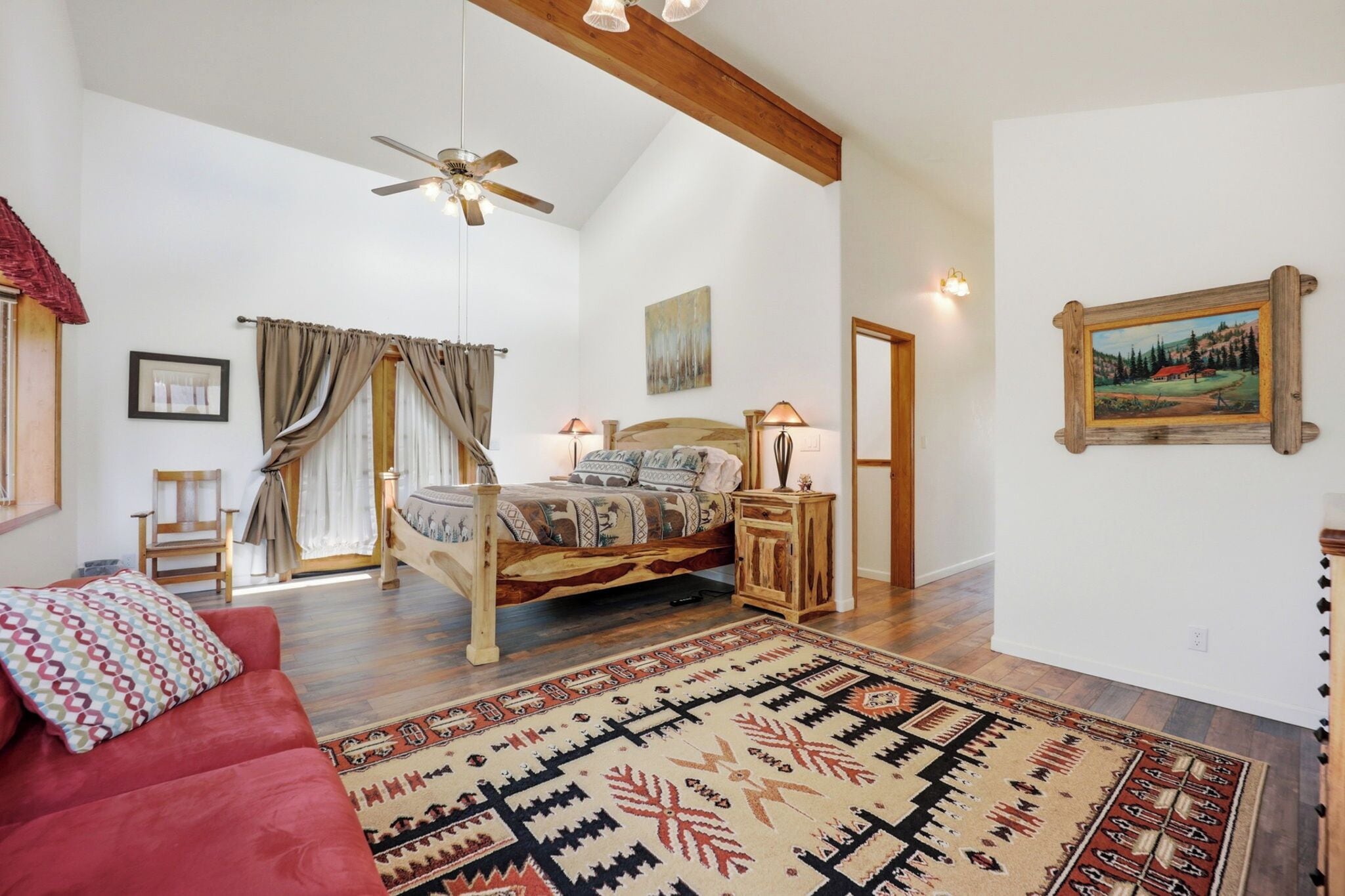 A spacious bedroom with a large bed, a ceiling fan, a wooden headboard, a nightstand, a red sofa, and a patterned rug.