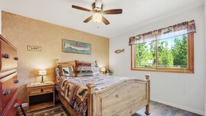 A bedroom with a wooden bed, a ceiling fan, a nightstand, a dresser, a rug, a fish wall art, and a window with a view of trees. - Large Home on 1 Acre: 2 Mi to Grand Lake Village (Grand Lake)