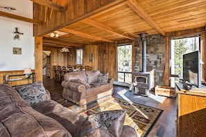 A cozy cabin interior with a wooden ceiling, a stone fireplace, a large sofa, and a dining area.
