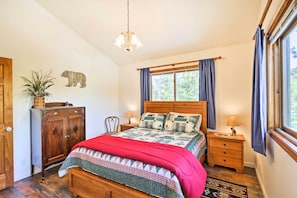 A bedroom with a wooden bed, a nightstand, a dresser, a chandelier, a window with curtains, and a wall-mounted bear decoration.