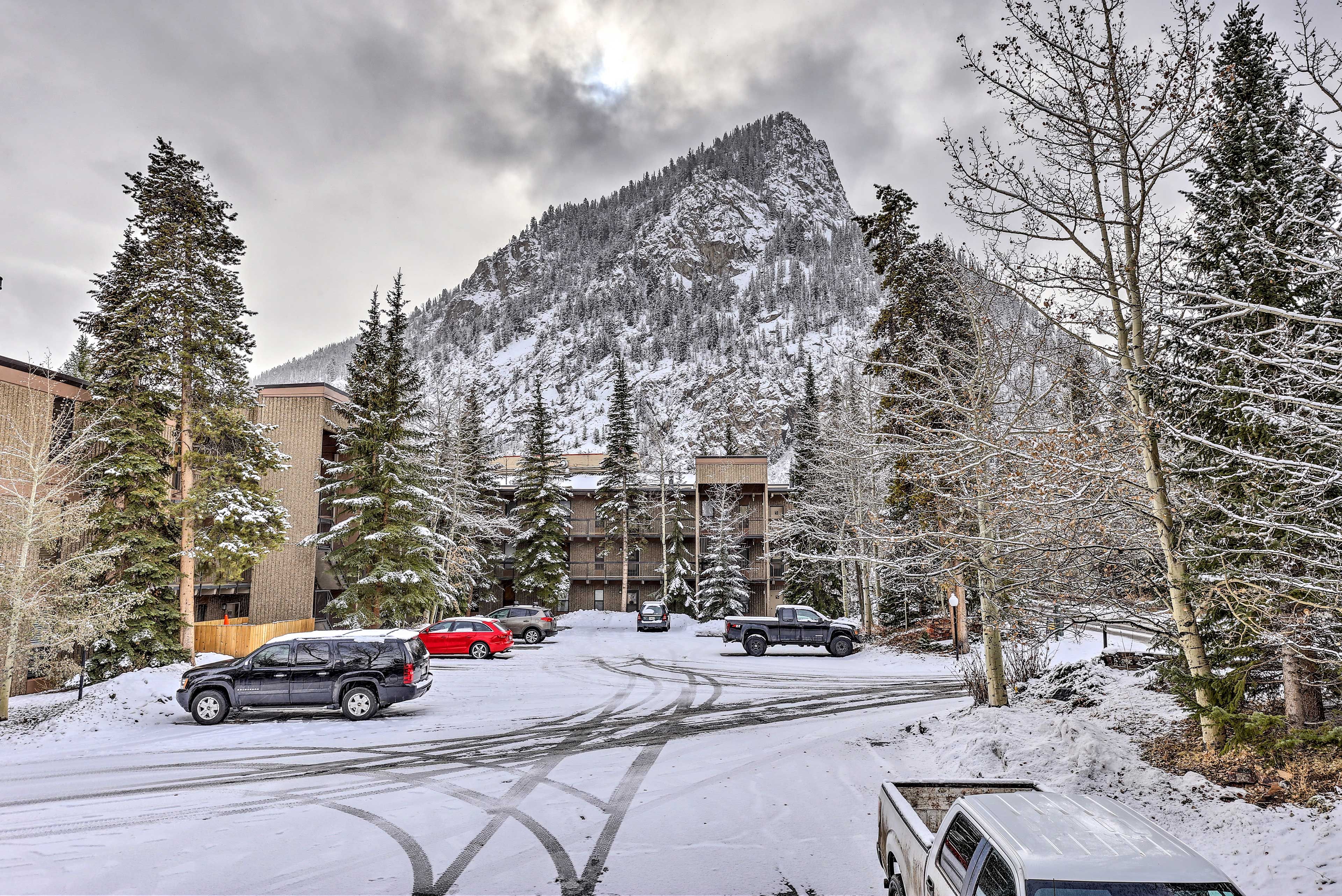 A snowy parking lot with vehicles, a mountain in the background, and a building with multiple floors.
