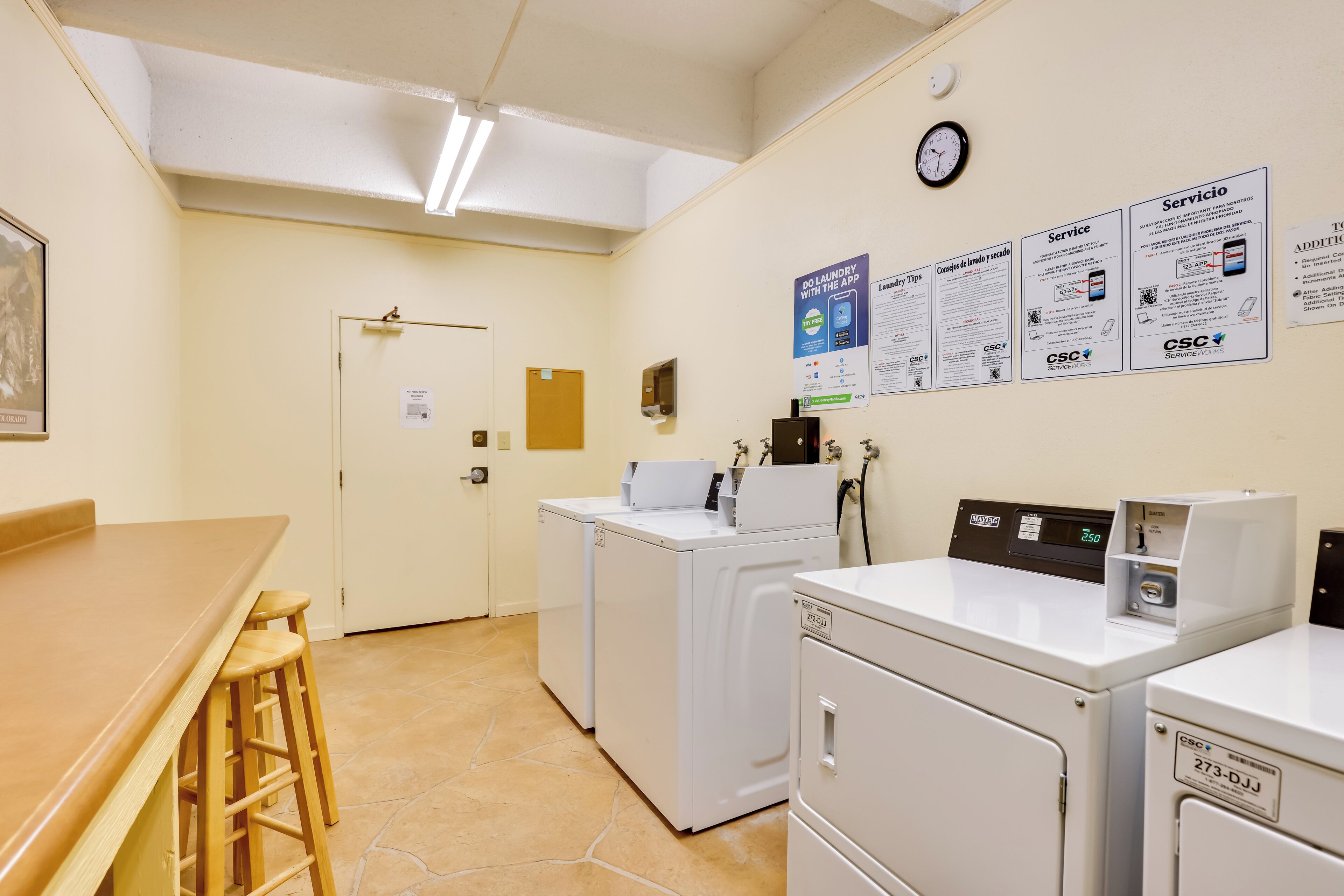 A laundry room with a washing machine, dryer, and a sink. There are stools and a table, and a clock on the wall.