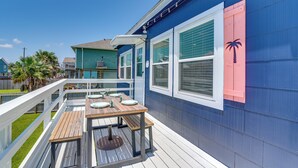 A deck with a wooden table and chairs, a white railing, and a blue building with a pink shutter.
