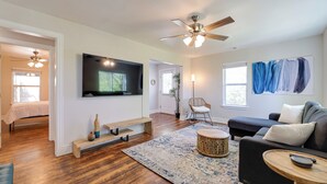 A spacious living room with hardwood flooring, a ceiling fan, a flat-screen TV, a dark-colored sofa, a round wooden coffee table, and a patterned rug. - Fayetteville Retreat w/ Yard - Walk to Campus! (Fayetteville)