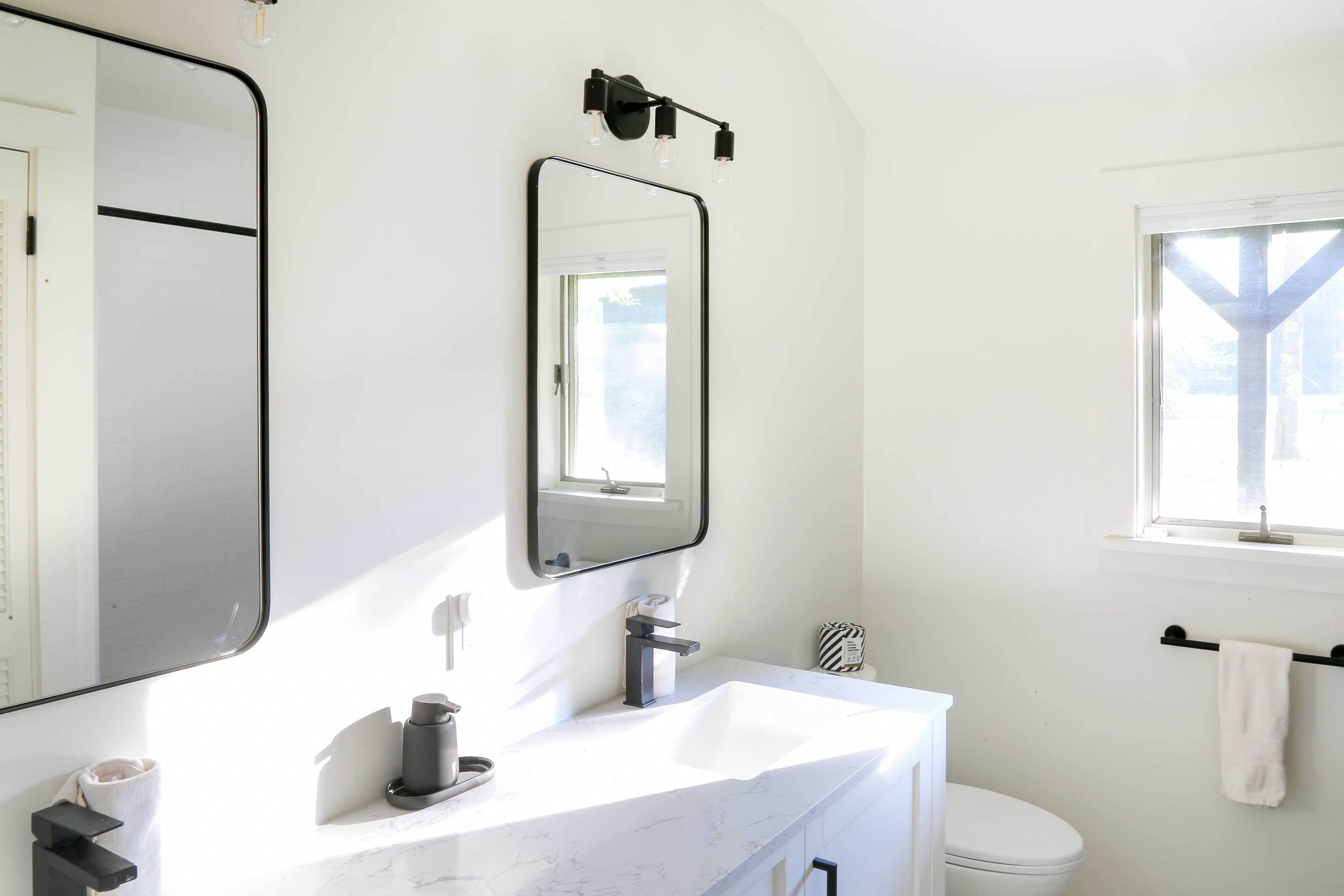 A modern bathroom with a white sink, black faucet, and a large mirror above it. There is a towel rack with a white towel hanging on it, and a window providing natural light.