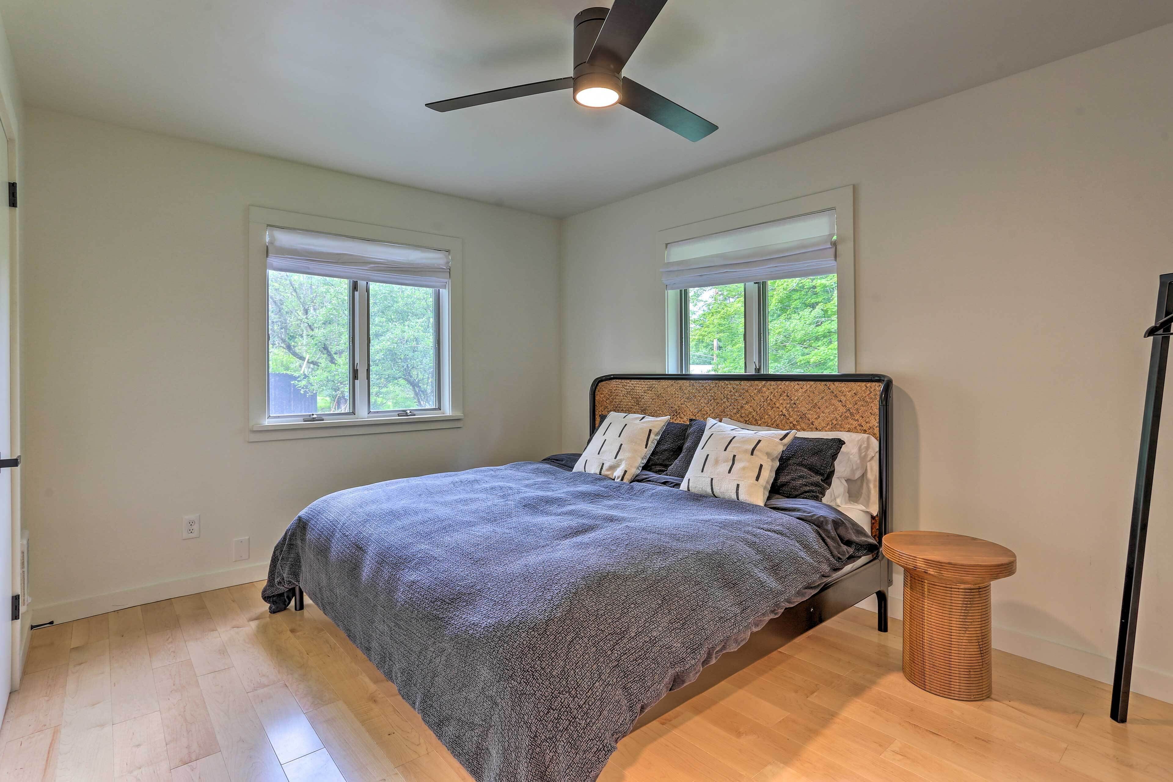 A bedroom with a bed, a ceiling fan, two windows, and a small wooden stool.