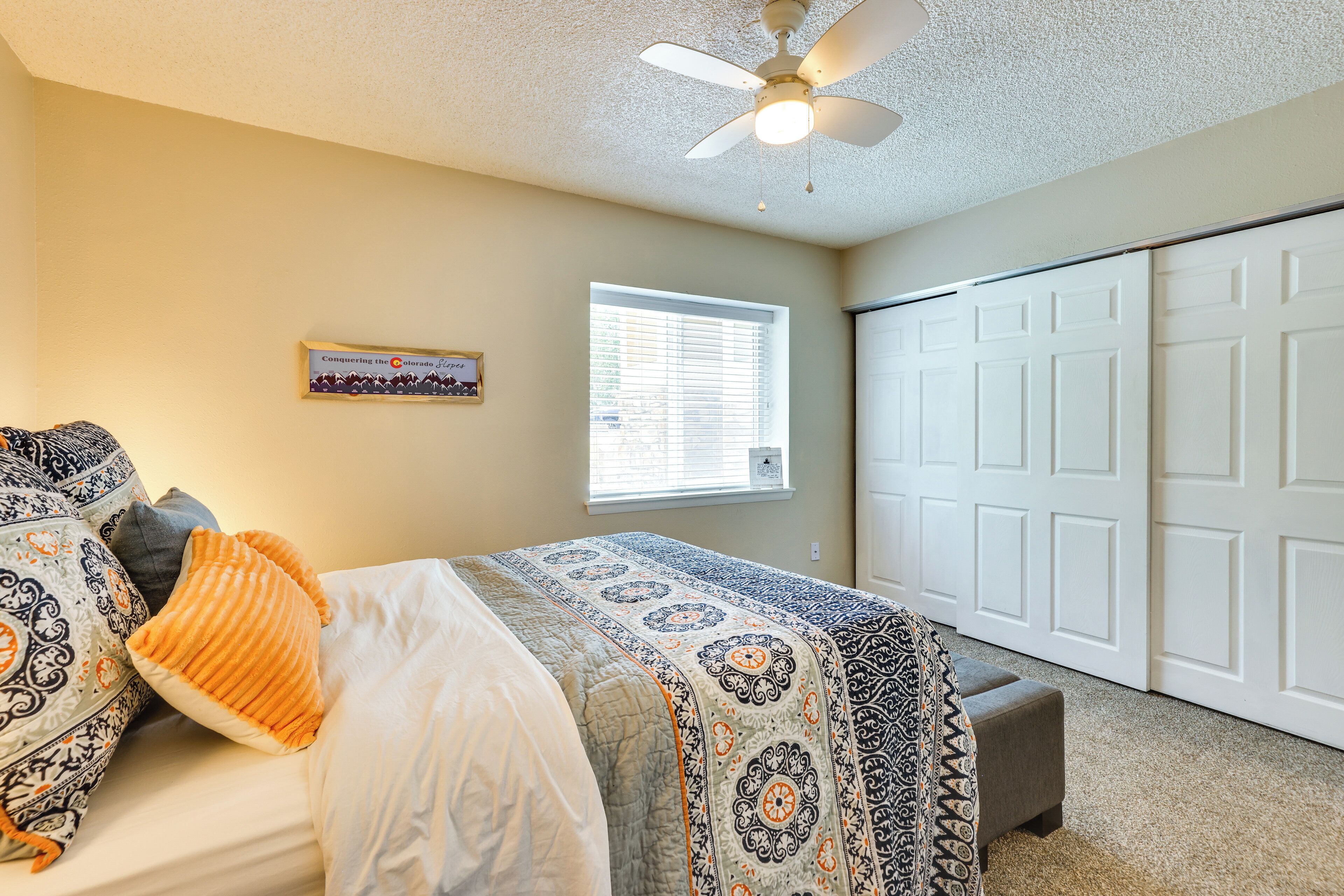 A bedroom with a bed, a ceiling fan, a window with blinds, a door, and a framed picture on the wall.
