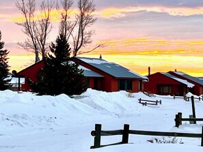 A row of red cabins with snow-covered roofs, surrounded by a wooden fence, set against a sunset sky. - Knapsack- Newly updated mountain cabin! (Clark)