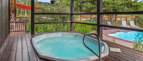 A wooden deck with a hot tub, outdoor seating, and a view of a swimming pool.