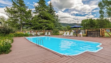 A rectangular swimming pool with a wooden deck surrounding it, equipped with lounge chairs and a covered seating area.