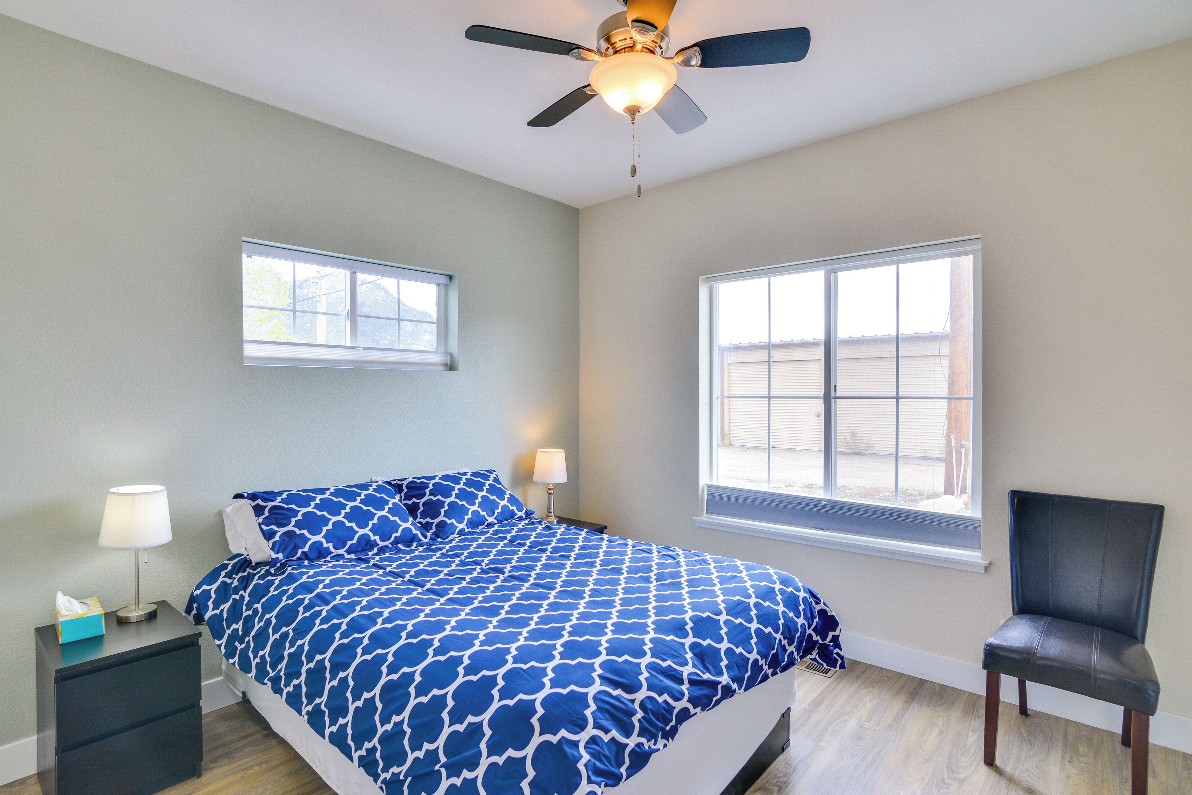 A bedroom with a bed, two windows, a ceiling fan, a nightstand, and a chair.