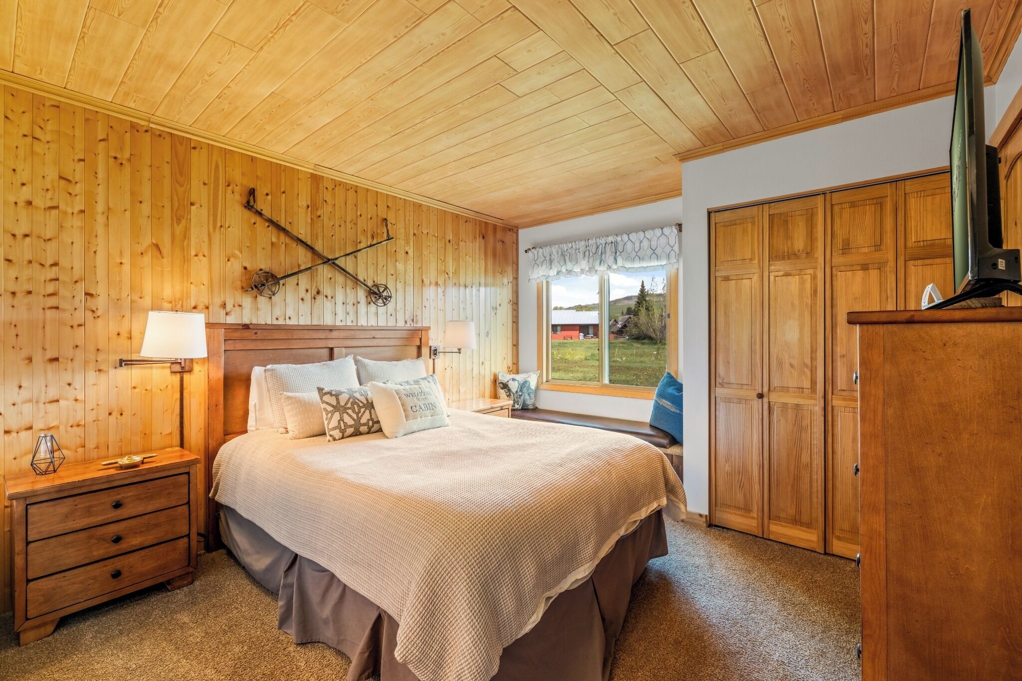Un dormitorio con techo de madera, una cama con cubrecama blanco, una mesita de noche, una lámpara y una ventana con vistas al exterior.