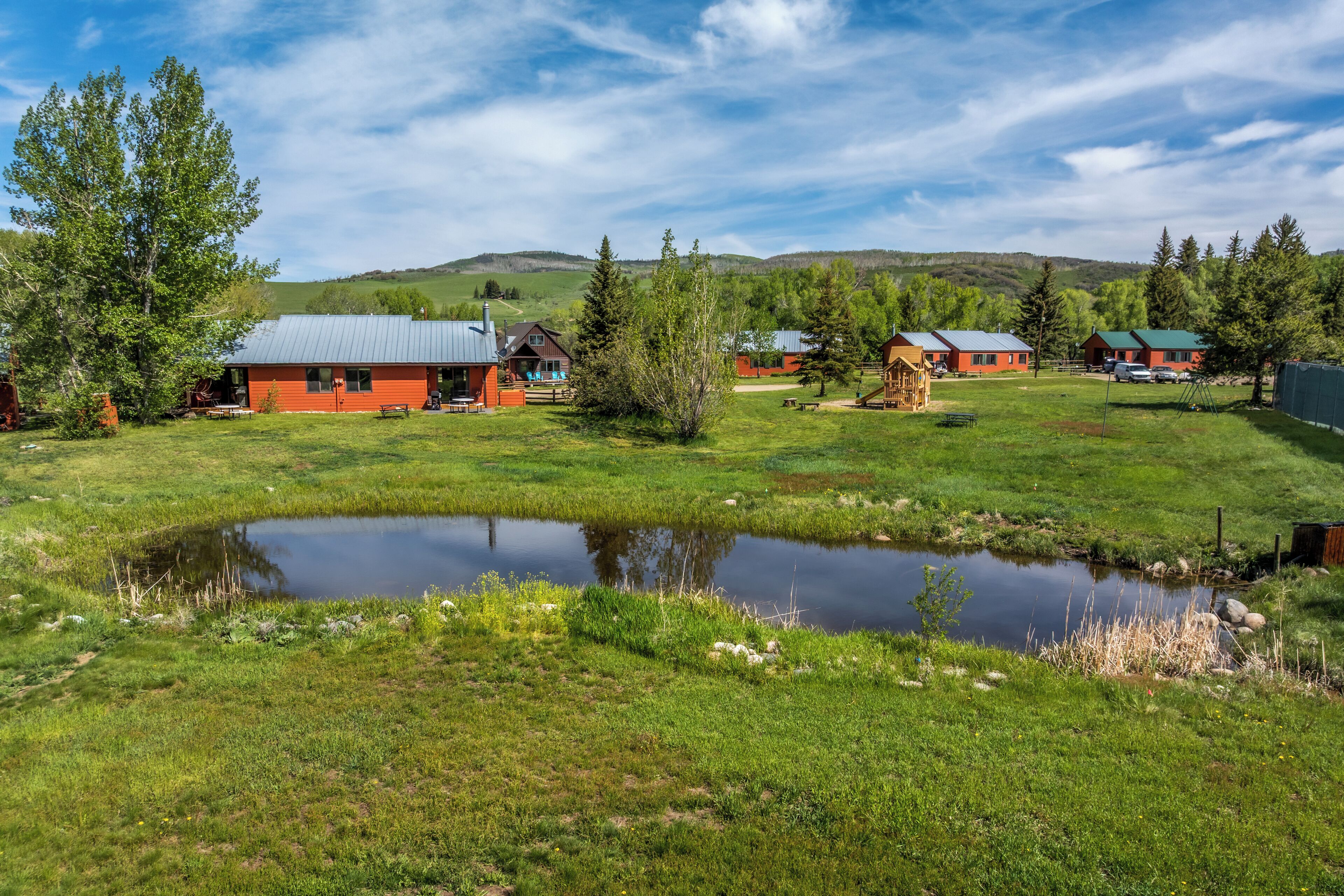 A rural setting with a pond, green fields, and several cabins with metal roofs.
