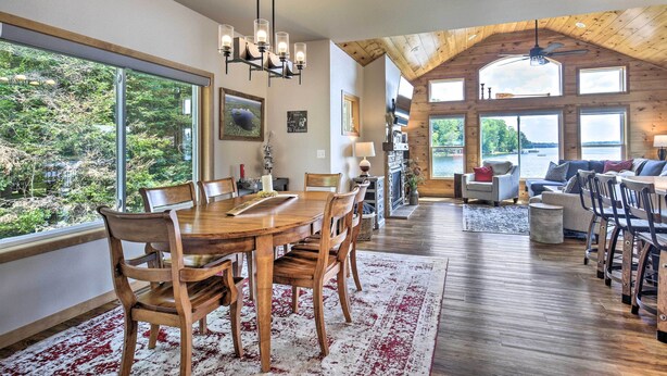 A spacious dining area with a large wooden table and chairs, a chandelier, and large windows offering a view of the outdoors. - Spacious Lakeside Family Home on Big Bearskin Lake (Harshaw)