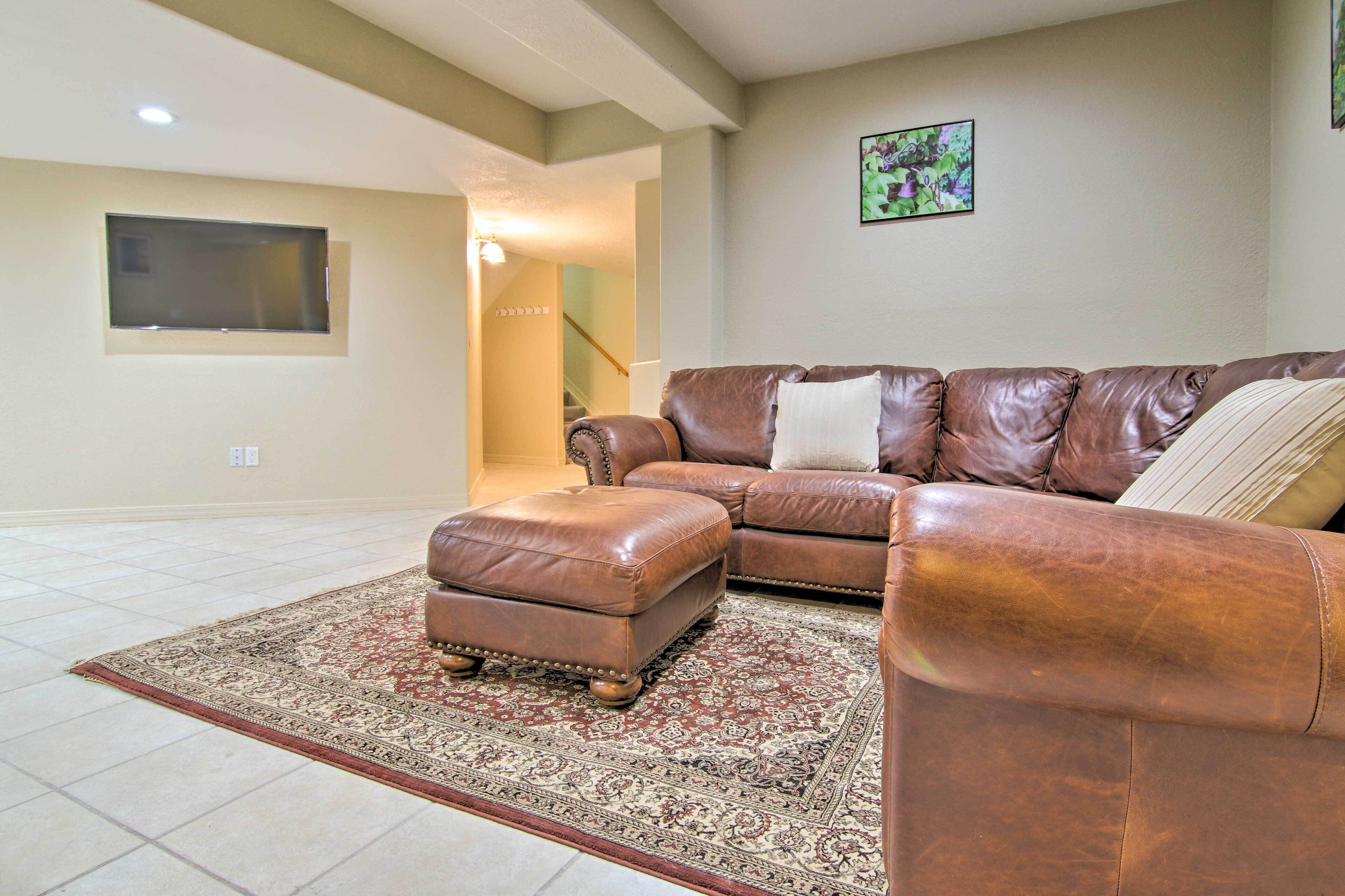 A spacious living room with a brown leather sectional sofa, a matching ottoman, and a patterned rug.