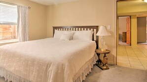 A neatly made bed with a white bedspread and pillows, a bedside lamp, and a view of a wooden door leading to another room. - CO Springs Apartment < 6 Mi to Garden of the Gods! (Colorado Springs)