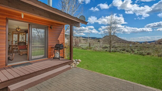 A wooden cabin with a patio, a sliding glass door leading to an interior with a dining area, and a view of a grassy field and mountains. - Fish Hook - Cozy cabin tucked in the mountains (Clark)