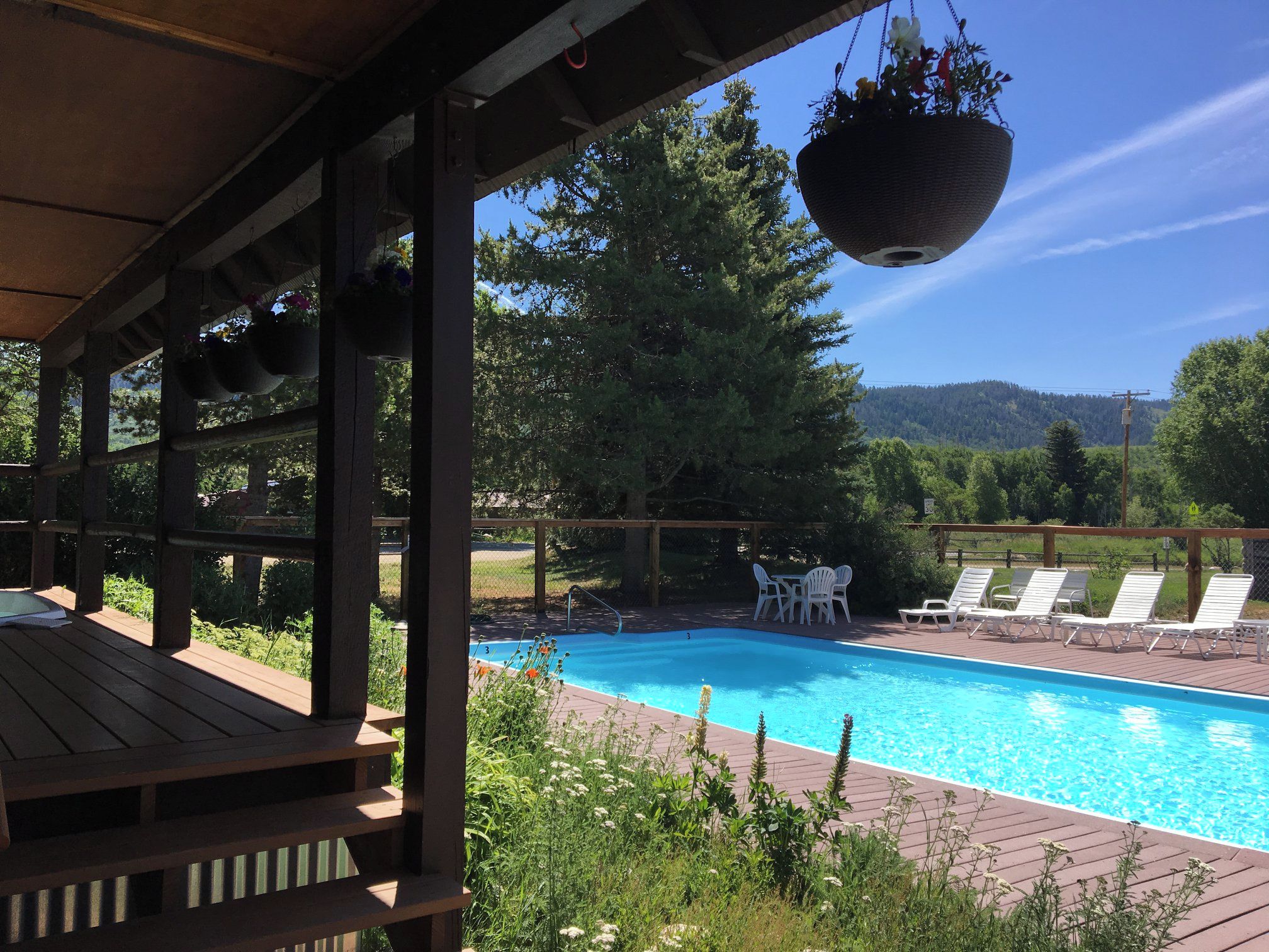 Terraza de madera con piscina, sillas de descanso blancas y cestas de flores colgantes.