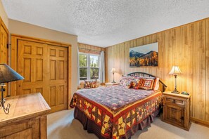 A bedroom with a large bed, two bedside tables, a wooden wardrobe, a window with a view of trees, and a painting on the wall. - Fish Hook - Cozy cabin tucked in the mountains (Clark)