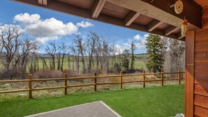 A covered outdoor area with a wooden fence, a brick patio, and a view of a grassy landscape with trees and hills. - Fish Hook - Cozy cabin tucked in the mountains (Clark)