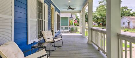A porch with two cushioned chairs and a table, a ceiling fan, and a view of the street with houses and flags.