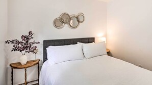 Una cama bien hecha con ropa de cama blanca, una mesita de noche con un florero y una planta, y un espejo montado en la pared sobre la cama.