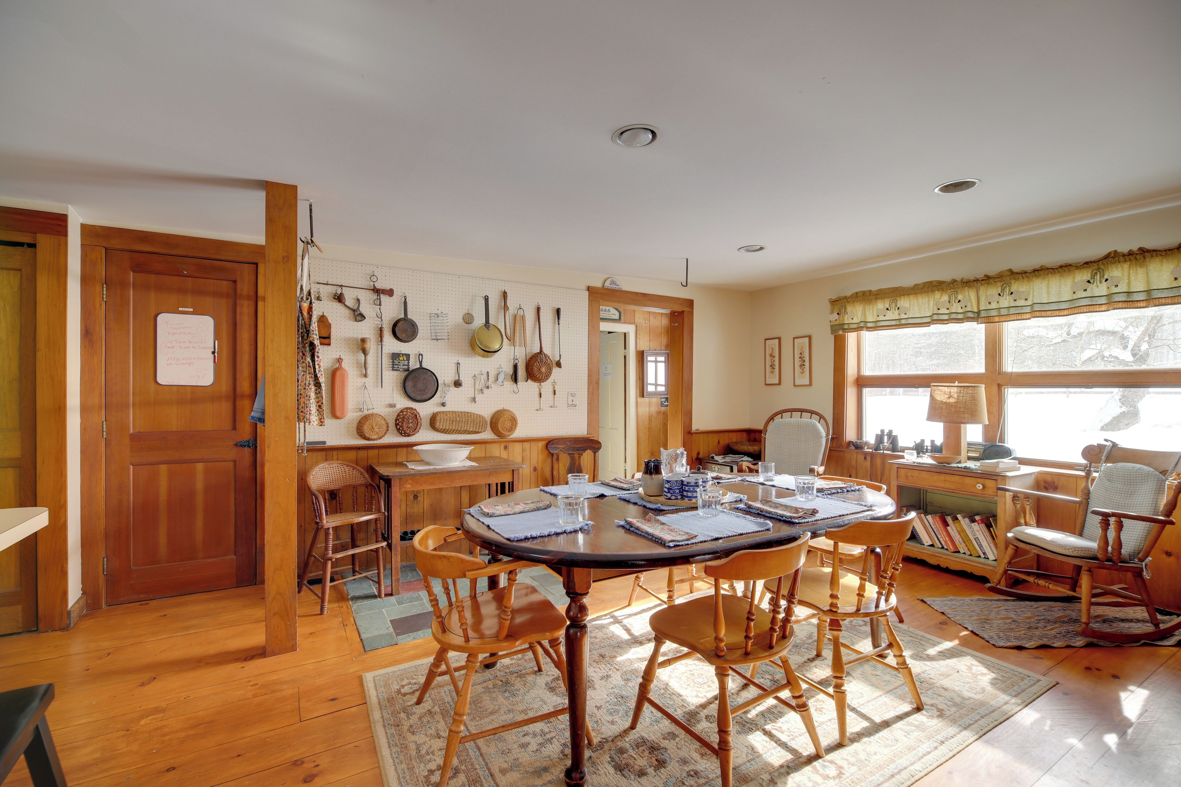 A dining area with a wooden table and chairs, a pegboard with kitchen utensils, and a window with curtains.