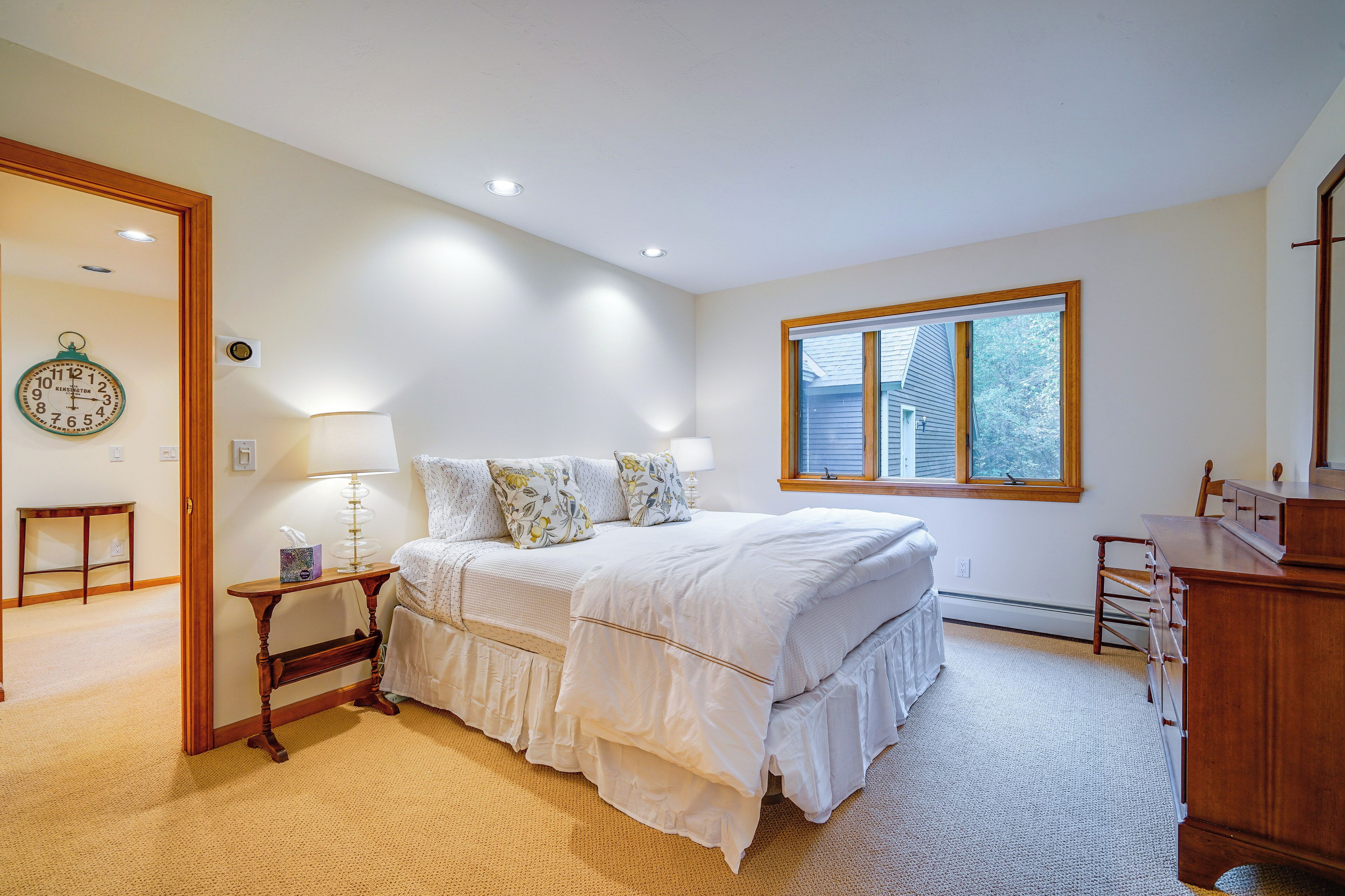 A bedroom with a bed, bedside table, lamp, dresser, and a window with a view of a house.