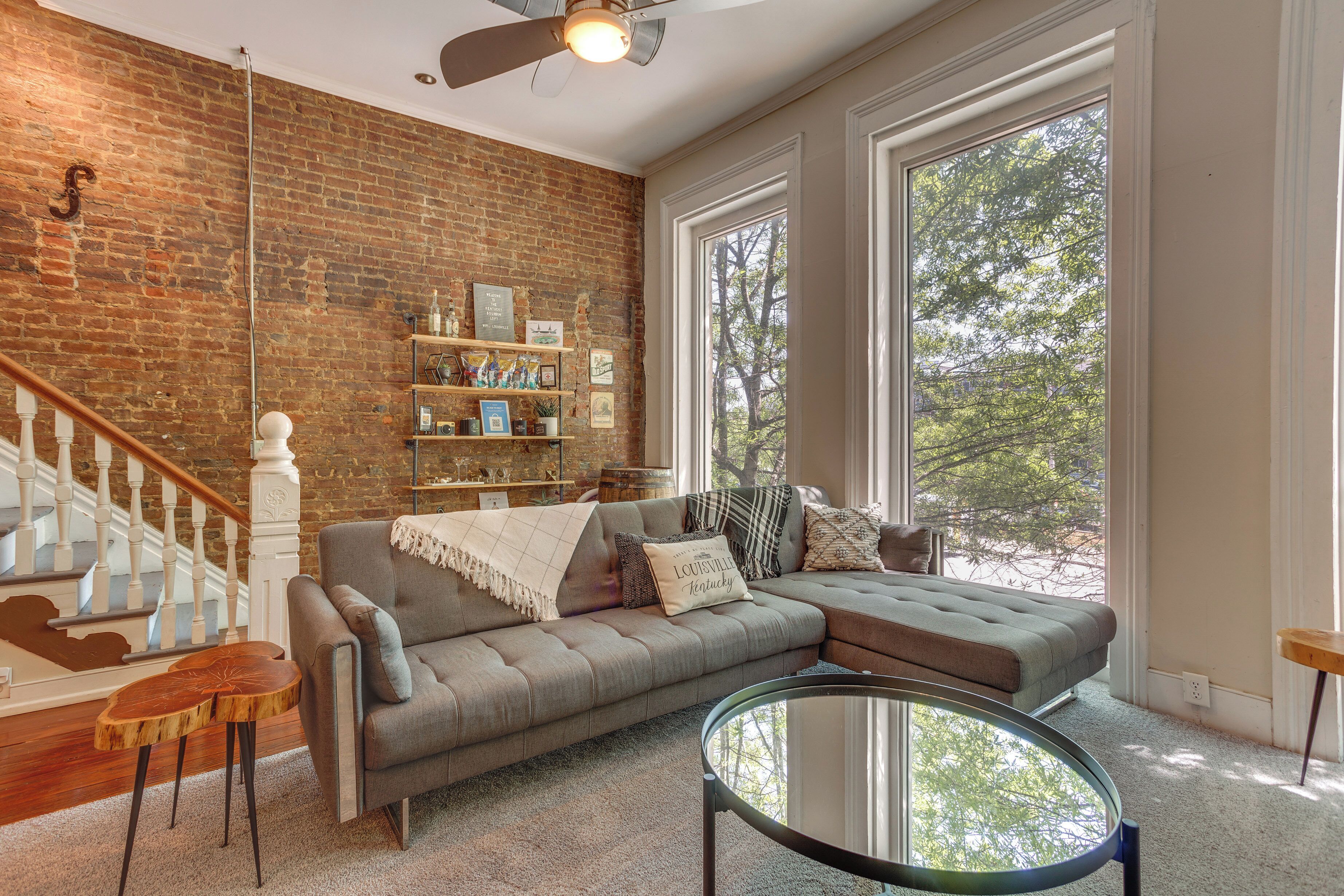 A living room with a brick wall, a gray sectional sofa, a glass coffee table, and a wooden staircase.