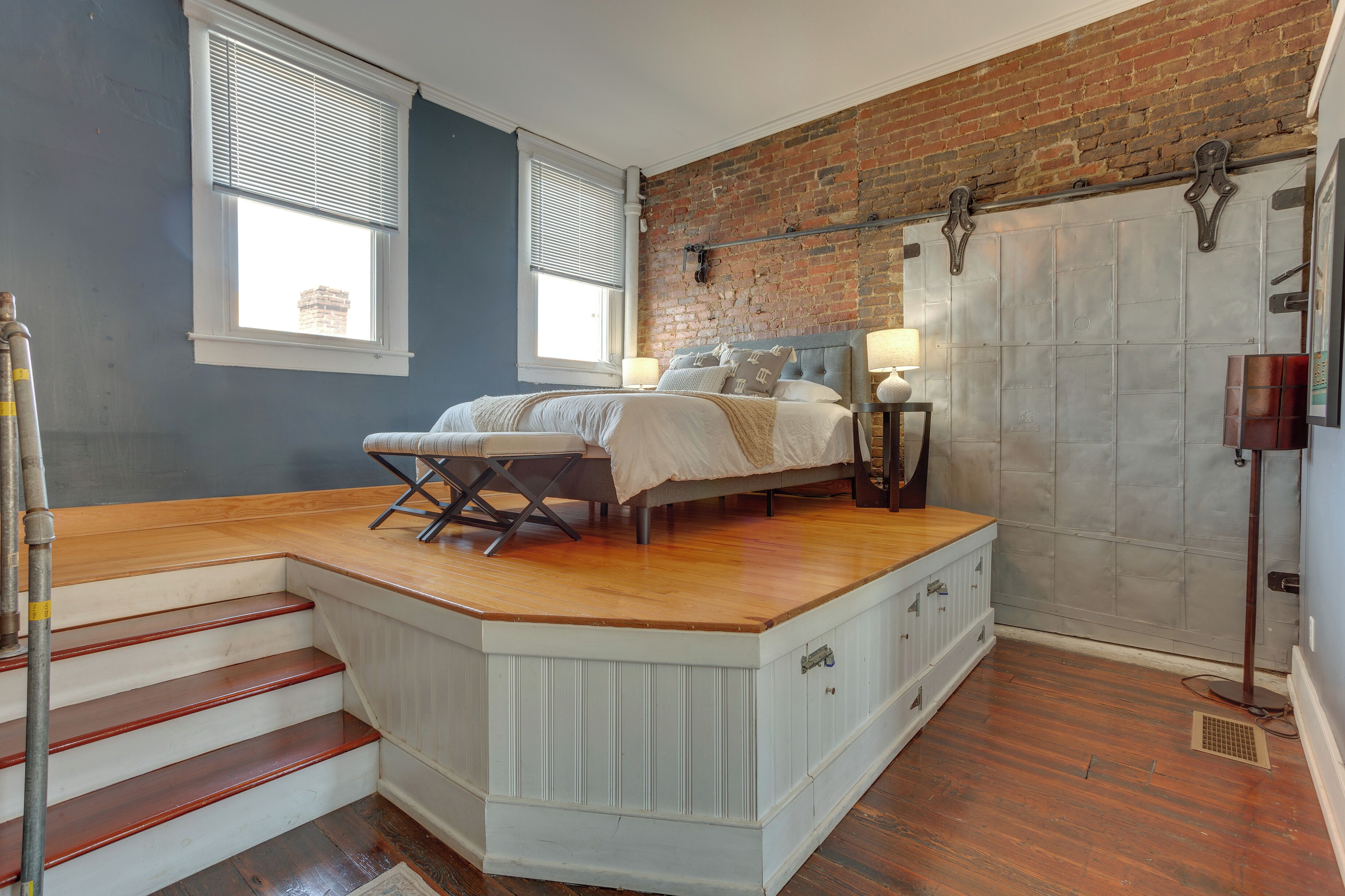 A bedroom with a bed, a nightstand, a lamp, a window, and a brick wall.