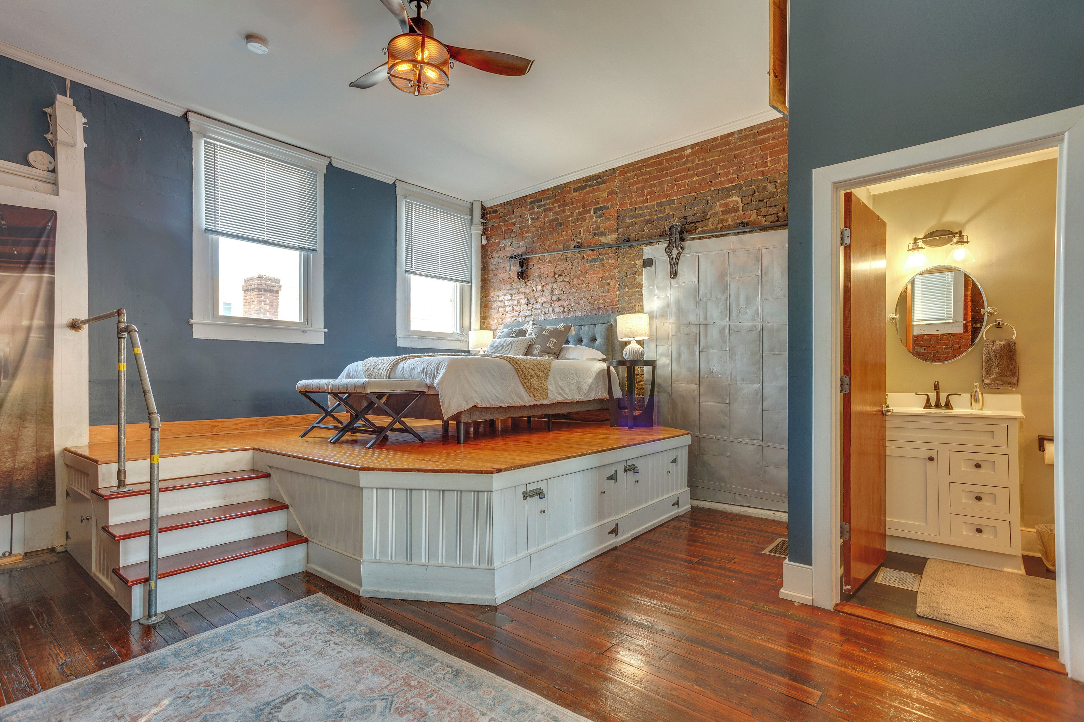 A bedroom with a loft bed, a brick wall, a wooden staircase, and a bathroom with a vanity and mirror.