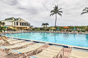 A pool area with lounge chairs and umbrellas, a building with a light-colored facade, and palm trees. - Hutchinson Island Condo: Community Amenities! (Fort Pierce)