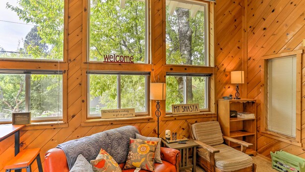 A cozy cabin interior with a leather sofa, a wooden chair, a small table, and a bookshelf. Large windows offer views of trees and a sign that reads "welcome". - Cozy Studio Near Hiking & Skiing, Walk to Downtown (Mount Shasta)