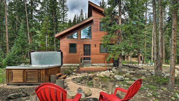 Eine moderne Holzblockhütte mit Whirlpool, zwei roten Adirondack-Stühlen und einer Terrasse mit Blick auf einen bewaldeten Bereich.