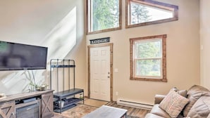 Ein Wohnzimmer mit Fernseher, einer Couch, einem Couchtisch und einem Kamin. Es gibt Fenster und eine Tür mit der Aufschrift 'Lodge'.
