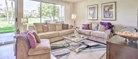 A spacious living room with two sofas, a coffee table, and a ceiling fan. Large windows provide a view of the outdoors.
