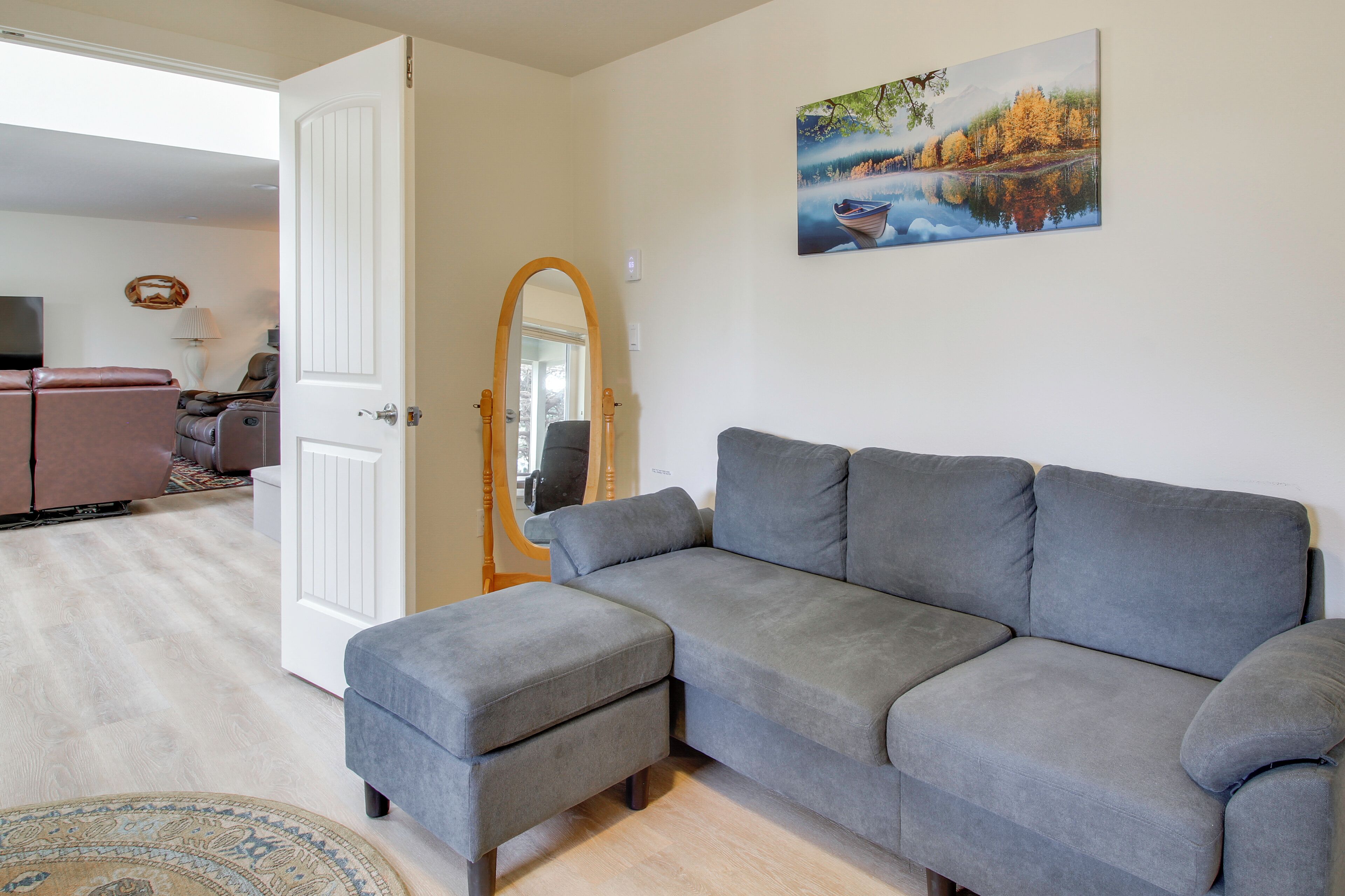 A living room with a grey sectional sofa, a small ottoman, a round rug, a painting of a lake, and a wooden-framed mirror.