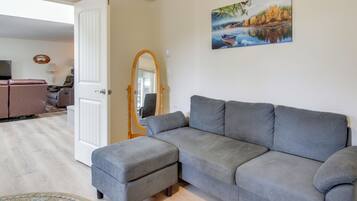 A living room with a grey sectional sofa, a small ottoman, a round rug, a painting of a lake, and a wooden-framed mirror.