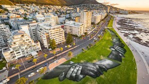 Paisagem urbana com uma montanha imponente ao fundo, uma estrada com veículos e um grande mural de mãos dadas.