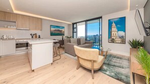 Sala de estar moderna com sofá, poltrona, mesa de centro e área de cozinha com ilha branca. Duas janelas amplas oferecem vista da cidade.