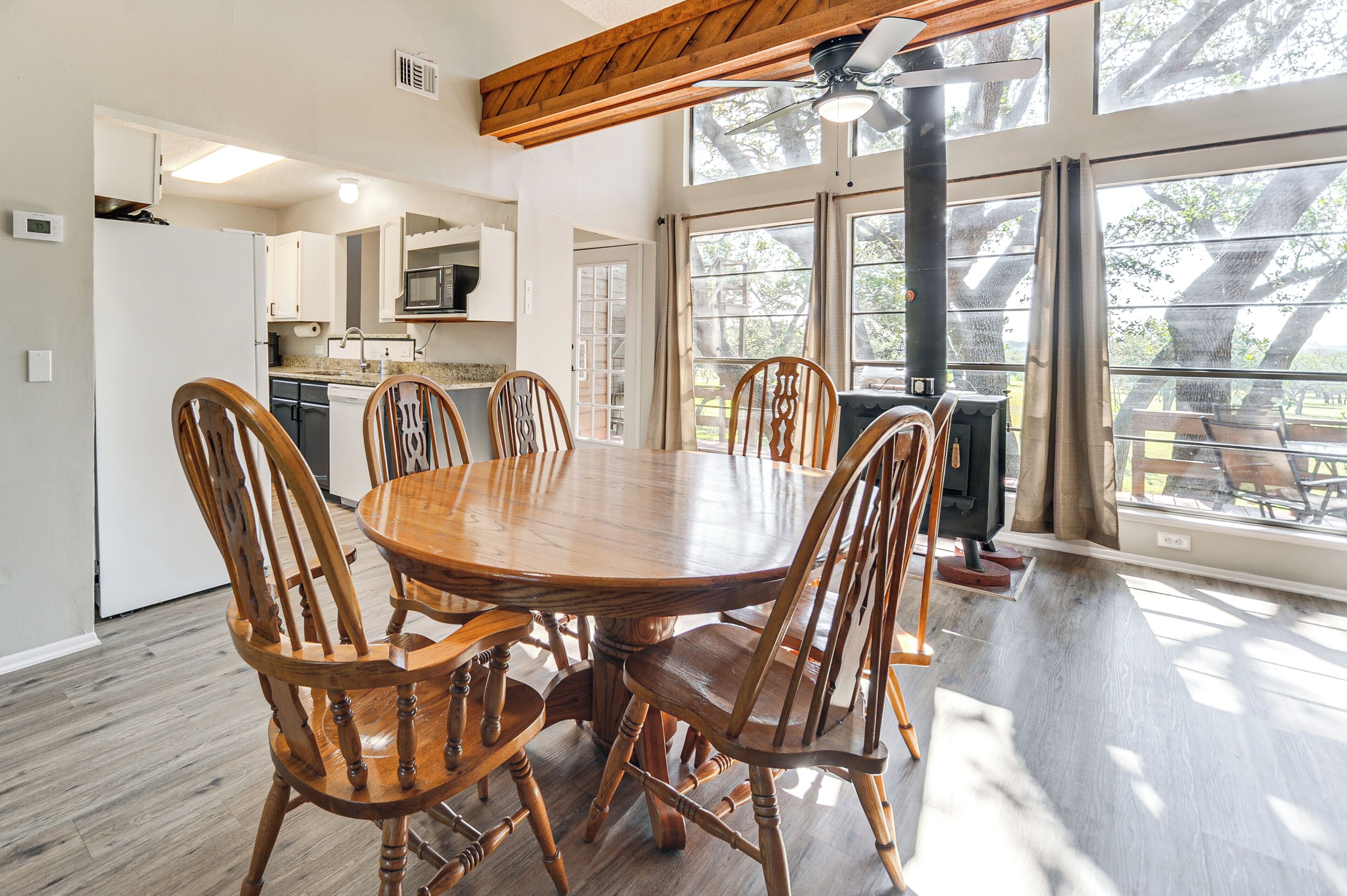 Un'ampia sala da pranzo con tavolo e sedie in legno, grandi finestre che si affacciano sugli alberi e una cucina sullo sfondo.