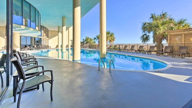 Un área de piscina con sillones reclinables, una estructura de cristal y palmeras.