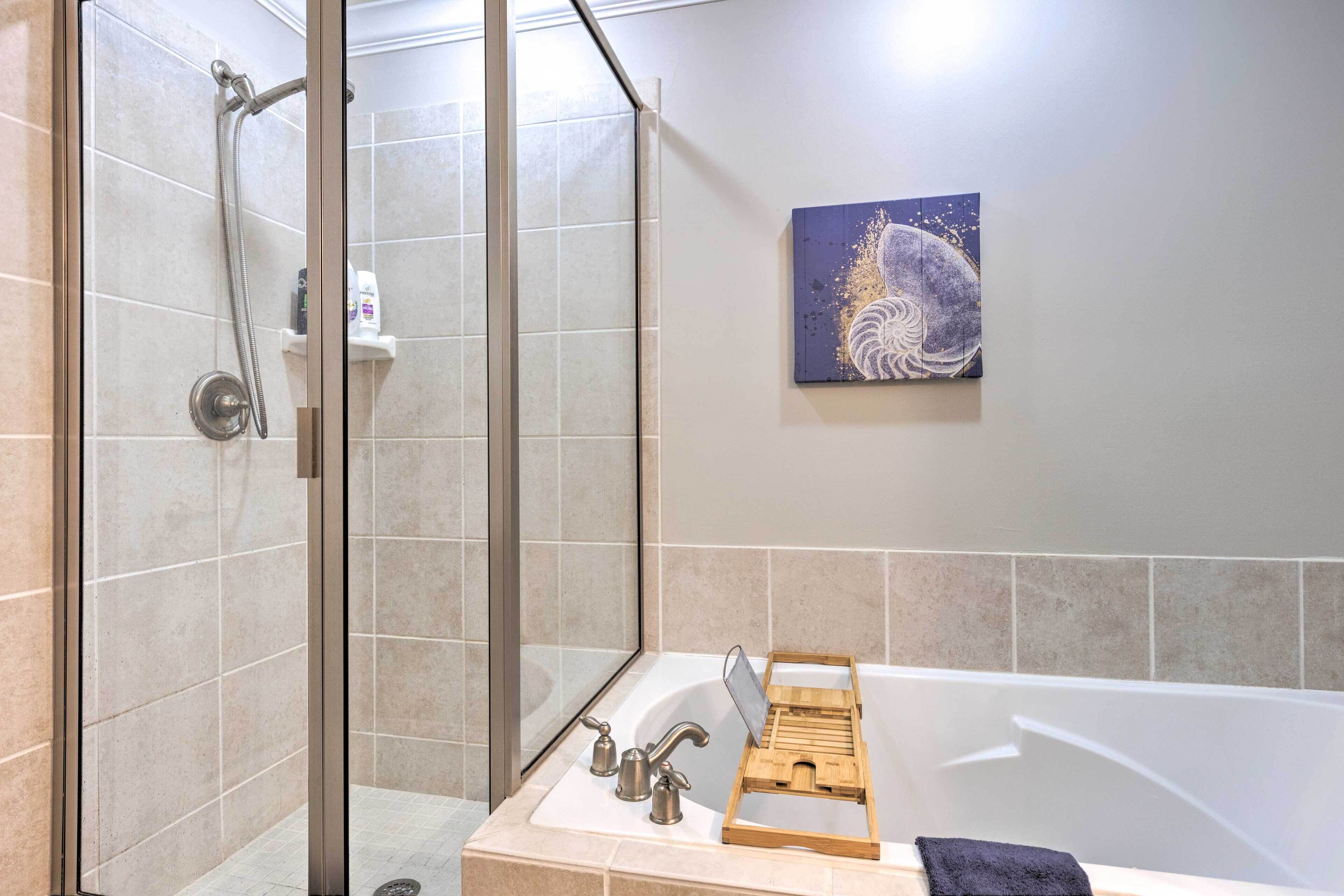 A modern bathroom with a glass-enclosed shower, a bathtub with a wooden tray, and a wall-mounted artwork.