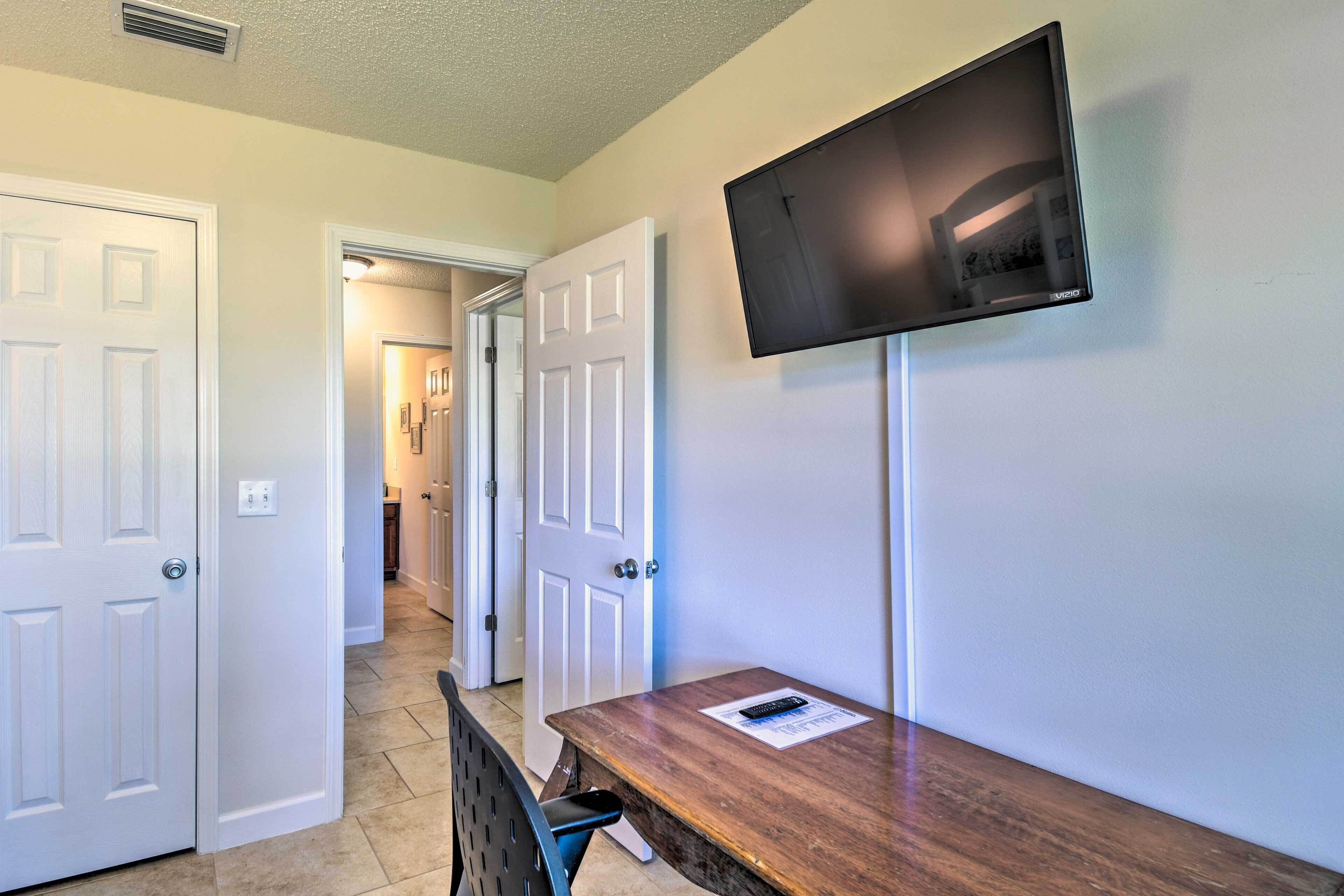 A room with a wooden desk, a mounted flat-screen TV, and a door leading to a hallway.