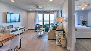 A spacious living area with a wooden dining table, a TV mounted on the wall, and a balcony with a view. - Ocean-view Balcony: Family Gem on Seawall Blvd! (Galveston)