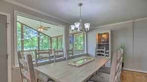A dining area with a large wooden table, chairs, a chandelier, and a view of the outdoors through large windows. - The Lake Escape: Boat Dock, Fire Pit, Kayak! (Cropwell)